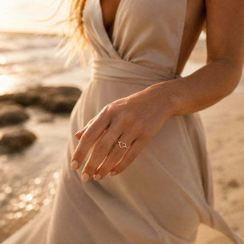 Blonde Frau in einem schicken Kleid präsentiert den Rosalie Ring Gold mit offener Herzform am Finger beim Gehen am Strand in warmem Abendlicht.