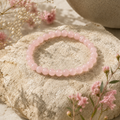 Rosenquarz-Natursteinarmband mit 6 mm großen, hellrosa Perlen auf einer natürlichen, hellen Steinoberfläche mit zarten rosa und weißen Blüten.