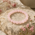 Moderne Frau trägt das Rosenquarz-Natursteinarmband mit 8 mm großen, rosafarbenen Perlen am Handgelenk auf einer hellen Terrasse am See mit rosa Blumen.