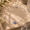 Elegante Safira Fußkette in Silber mit funkelndem Nazar Auge als traditionellem Schutzsymbol und Glücksbringer. Hochwertig inszeniert auf feinem Sand mit mediterranen Lichtreflexionen, silbernen Ornamenten und sommerlicher Atmosphäre. Filigraner Schmuck für stilvolle Alltagslooks, Urlaub und besondere Anlässe.