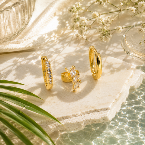 Elegantes Produktbild goldfarbener Ohrringe mit funkelnden Zirkonia-Steinen auf heller Natursteinplatte, umgeben von grünen Palmenblättern, Wasserreflexionen und sommerlicher Resort-Atmosphäre in luxuriöser AVRE Studios Ästhetik.