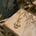 Goldfarbene Hand der Fatima Halskette mit blauem Nazar-Auge und funkelnden Details auf hellem Steinpodest. Elegante Schmuckinszenierung mit grünen Blättern, warmem Licht und orientalischen Dekorelementen im AVRE Studios Stil.
