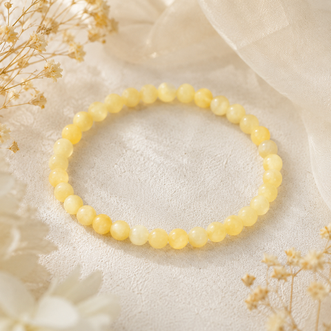 Yellow Calcite Jade Natursteinarmband mit 6 mm großen, leicht marmorierten Perlen in zarten Gelb- und Cremetönen auf hellem Stoff mit dezenten Trockenblumen präsentiert.