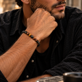 Mann trägt das KAIO Tigerauge & Lavastein Armband mit fünf kleinen Natursteinperlen und schwarzem geflochtenem Band.