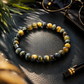 Tigerauge-Armband mit 6-mm-Perlen in Blau-, Grau-, Gold- und Brauntönen auf dunklem, eleganten Hintergrund mit warmem Licht und stilvollen Accessoires.