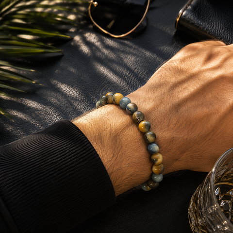 Nahaufnahme eines Herrenarms mit Tigerauge-Armband in Blau-, Grau-, Gold- und Brauntönen vor dunklem, stilvollem Hintergrund mit warmem Licht und eleganten Details.