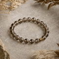 Facettiertes Rauchquarz-Armband mit 8 mm großen braunen Perlen und runder goldfarbener Akzentperle auf hellem Naturuntergrund mit Trockenblumen.